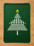 Green chunky knit blanket featuring a white abstract knit Christmas tree pattern with a yellow star on top, displayed flat on a light wood floor.