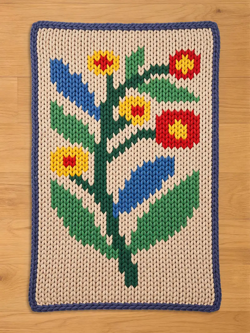 A chunky knit blanket featuring an abstract floral pattern with red, yellow, and blue flowers and green leaves on a beige background, framed by a dark blue border, lying flat on a wooden floor.
