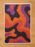 A chunky knit blanket featuring an abstract camo pattern in vibrant orange, dark grey, purple, and coral red, finished with a thick cream braided border, lying flat on a wooden floor