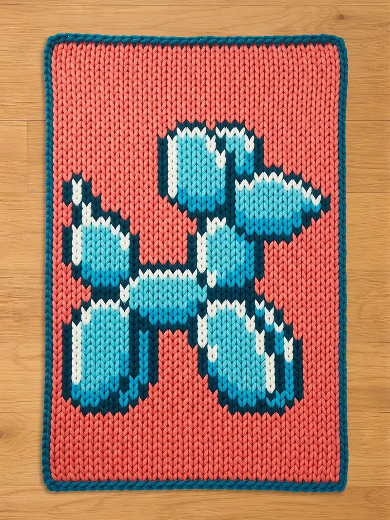 Chunky knit blanket featuring a pixelated blue balloon dog design on a coral pink background with a dark teal border, laid flat on a wooden floor.