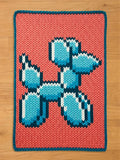 Chunky knit blanket featuring a pixelated blue balloon dog design on a coral pink background with a dark teal border, laid flat on a wooden floor.