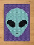 A knitted blanket with an alien face design in shades of purple, light blue, and black. The alien's head is light blue with black eyes and a small smile. The background is a darker purple