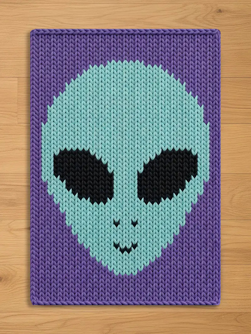 A knitted blanket with an alien face design in shades of purple, light blue, and black. The alien's head is light blue with black eyes and a small smile. The background is a darker purple