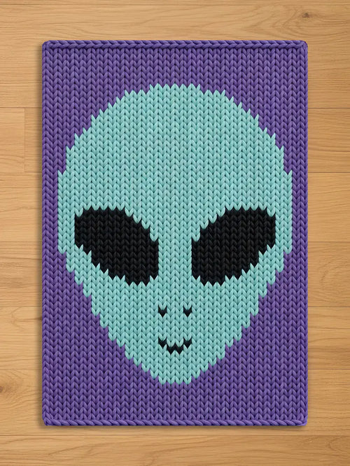 A knitted blanket with an alien face design in shades of purple, light blue, and black. The alien's head is light blue with black eyes and a small smile. The background is a darker purple