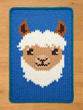 A blue knitted blanket featuring a llama face. The llama has a white woolly face, brown ears, and a tan muzzle. The blanket has a blue border and is set against a wooden floor.