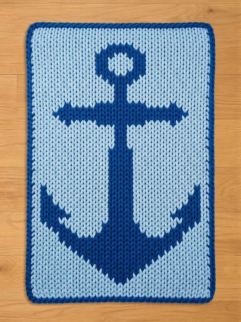 A rectangular, knitted blanket featuring a navy blue anchor design on a light blue background. The anchor is centered and detailed, with a dark blue border. The blanket is placed on a wooden surface.