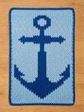 A rectangular, knitted blanket featuring a navy blue anchor design on a light blue background. The anchor is centered and detailed, with a dark blue border. The blanket is placed on a wooden surface.