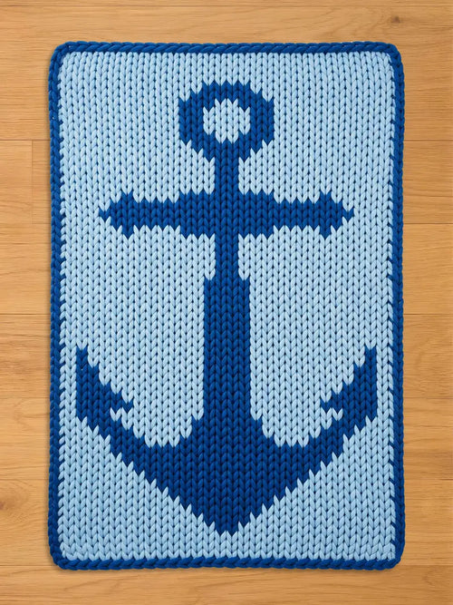 A rectangular, knitted blanket featuring a navy blue anchor design on a light blue background. The anchor is centered and detailed, with a dark blue border. The blanket is placed on a wooden surface.