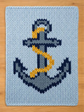 A light blue chunky knit blanket featuring a large, centered anchor design. The anchor is rendered in shades of dark navy blue, grey, and white, with a prominent yellow rope wrapped around the central shaft of the anchor structure.