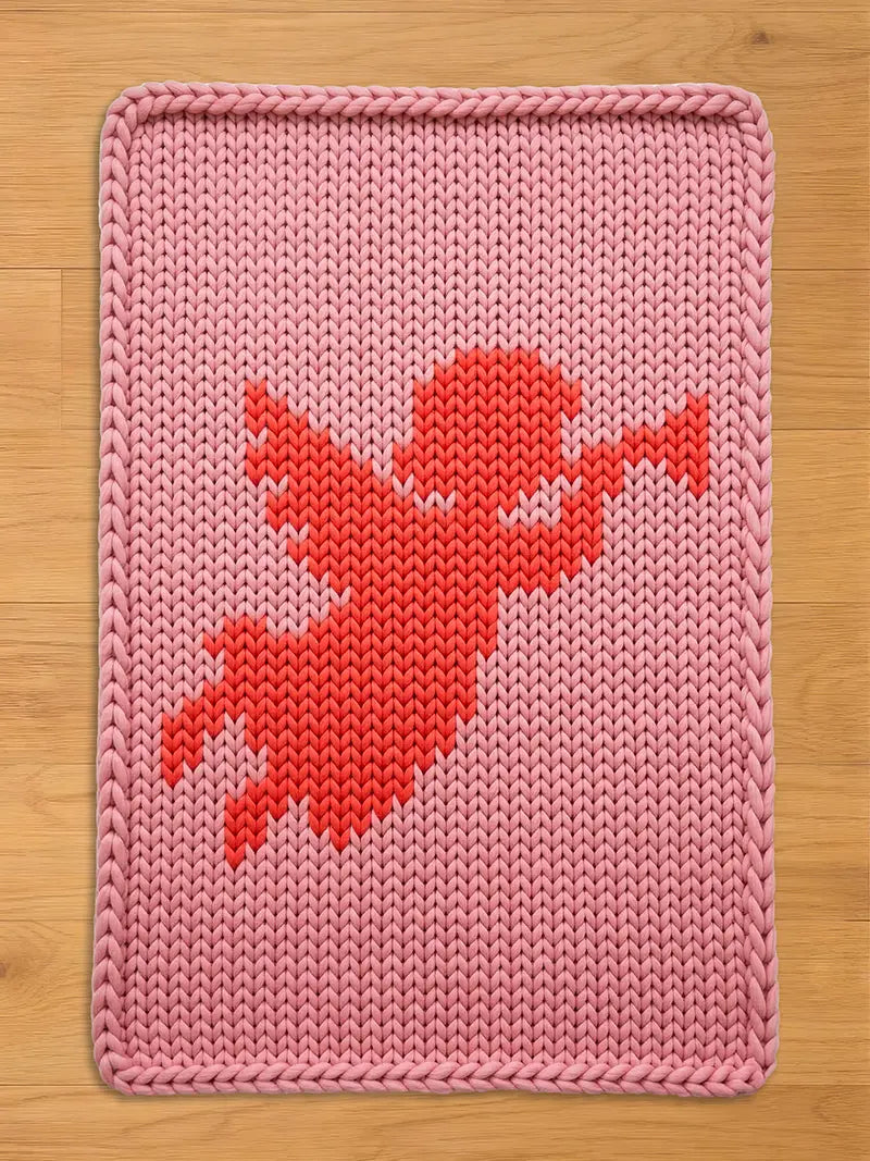 A pink knitted blanket with a red cupid design. The blanket has a rectangular shape with a thick, braided border. The cupid is depicted in a pixelated style, with outstretched arms and wings, against a light pink background.