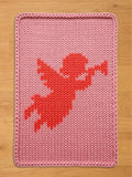 A pink knitted blanket with a red cupid design. The blanket has a rectangular shape with a thick, braided border. The cupid is depicted in a pixelated style, with outstretched arms and wings, against a light pink background.