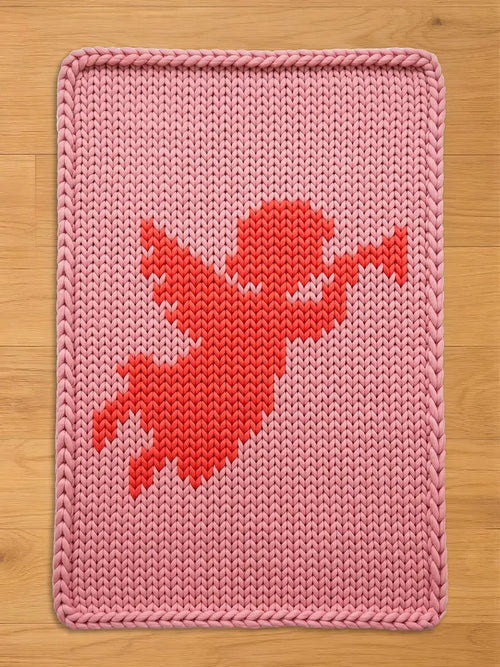 A pink knitted blanket with a red cupid design. The blanket has a rectangular shape with a thick, braided border. The cupid is depicted in a pixelated style, with outstretched arms and wings, against a light pink background.