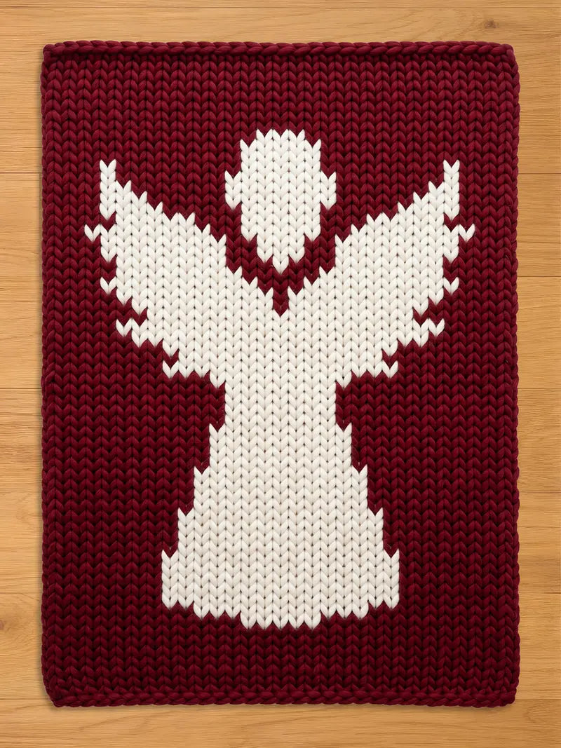 A rectangular knitted blanket featuring a large, white, stylized angel figure centered against a deep burgundy background. The piece is crafted with a chunky knit texture, showcasing a clear contrast between the white and dark red yarn.