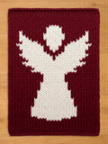 A rectangular knitted blanket featuring a large, white, stylized angel figure centered against a deep burgundy background. The piece is crafted with a chunky knit texture, showcasing a clear contrast between the white and dark red yarn.