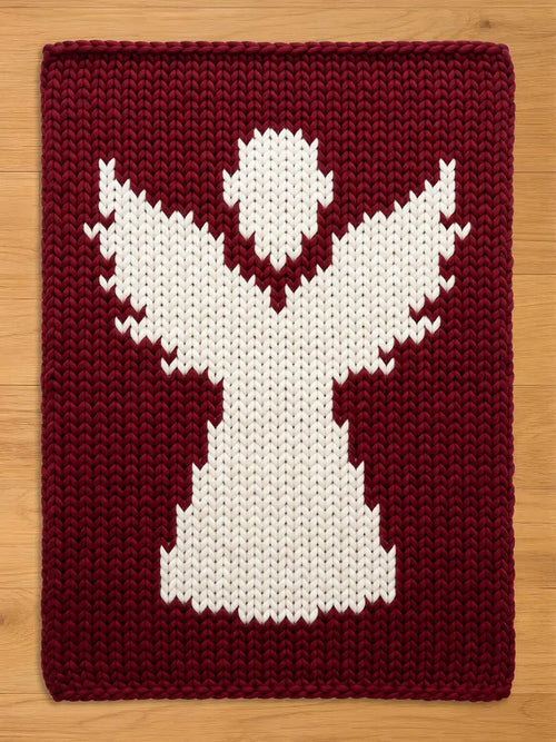 A rectangular knitted blanket featuring a large, white, stylized angel figure centered against a deep burgundy background. The piece is crafted with a chunky knit texture, showcasing a clear contrast between the white and dark red yarn.