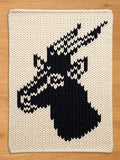 A square, off-white knitted throw blanket featuring a black silhouette of a deer head. The deer head is detailed with white chevron patterns. The blanket has a thick, textured appearance and a simple border.