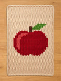 Chunky knit cream blanket featuring a large red apple pattern with a green leaf and brown stem, laid flat on a wooden floor.