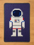 A chunky knitted blanket featuring a pixelated white astronaut design with red and blue accents on a dark purple background, laid flat on a wooden floor.