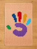A chunky knit peach blanket featuring a colorful, handprint design in the center with a purple palm and multi-colored fingers, laid flat on a light wood floor. Autism awareness symbol