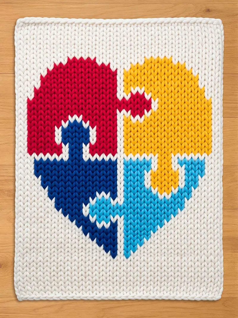 A square, knitted blanket featuring a heart design made of four puzzle pieces. The heart is divided into red, yellow, blue, and light blue sections, set against a white background. The blanket is made of thick yarn.
