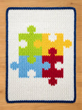 A white knitted blanket with a blue border. The blanket features a colorful puzzle design with pieces in green, yellow, red, and blue. The blanket is made of a soft, textured material.