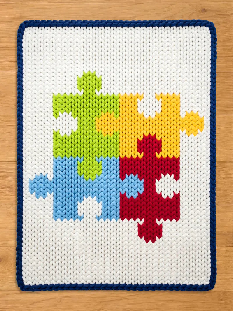 A white knitted blanket with a blue border. The blanket features a colorful puzzle design with pieces in green, yellow, red, and blue. The blanket is made of a soft, textured material.