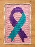 A chunky knit blanket featuring a large teal and purple awareness ribbon centered on a light pink background, finished with a cream-colored braided border, displayed flat on a wooden floor.