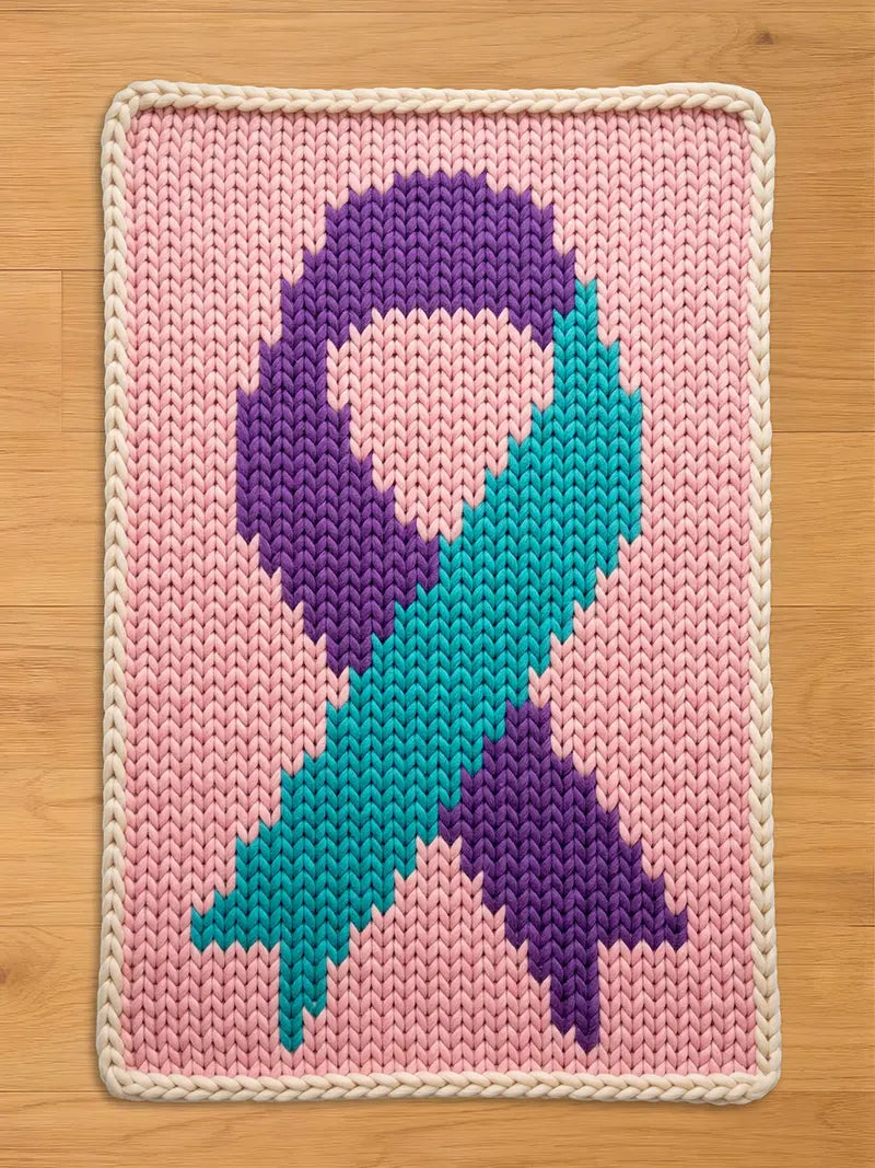 A chunky knit blanket featuring a large teal and purple awareness ribbon centered on a light pink background, finished with a cream-colored braided border, displayed flat on a wooden floor.
