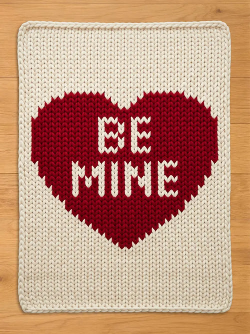 Chunky knit cream blanket featuring a large red heart in the center with the text "BE MINE" knitted inside