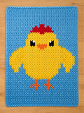 A knitted square blanket featuring a yellow chick design on a blue background. The chick has a red crest, black eyes, and orange beak and feet. The blanket is made of a thick, textured yarn, and is likely handmade.