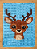 A knitted blanket featuring a pixelated deer design. The deer has brown antlers, a brown body, and a beige face. The background is a solid light blue color. The blanket is made of a soft, chunky knit material.