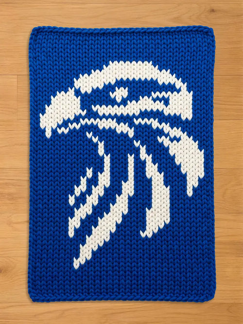 A knitted blue and white decorative square with a stylized design. The design features a white abstract eagle shape against a solid blue background. The item is made of a thick, textured yarn.