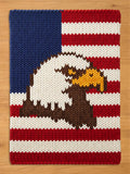 A rectangular knitted blanket featuring a patriotic design with a dark blue field in the upper left corner and horizontal red and white stripes. A large, detailed bald eagle with white head feathers and a yellow beak is centered.