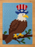 A rectangular knit blanket featuring a large bald eagle with a white head, brown body, and yellow beak, perched on a brown branch with green leaves. The eagle wears a tall top hat with red, white, and blue stripes and stars.