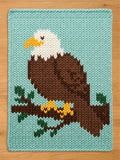 A rectangular knit blanket featuring a pixelated design of a bald eagle with a white head, yellow beak, and brown body, perched on a brown branch with green leaves against a light blue background, crafted with thick yarn stitches.