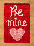 A vibrant red chunky knit blanket featuring the words "Be Mine" in a white cursive font above a large light pink knit heart, laid flat on a light wood floor