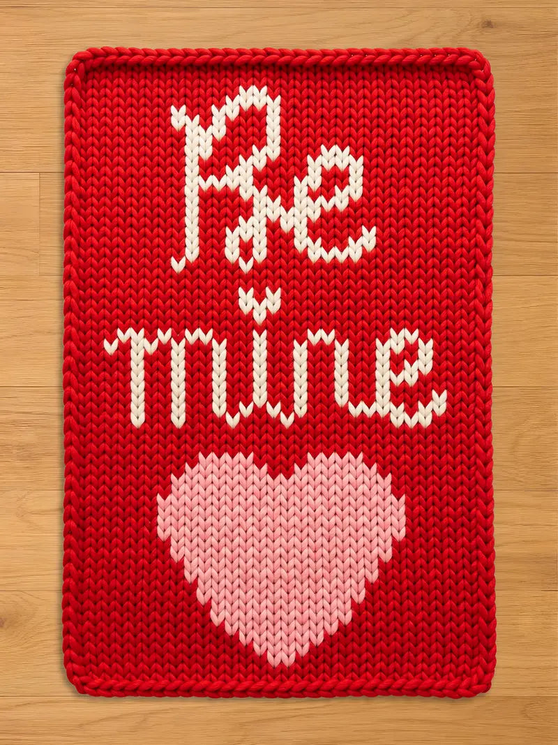 A vibrant red chunky knit blanket featuring the words "Be Mine" in a white cursive font above a large light pink knit heart, laid flat on a light wood floor