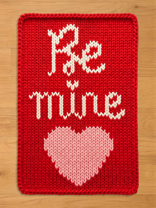 A vibrant red chunky knit blanket featuring the words "Be Mine" in a white cursive font above a large light pink knit heart, laid flat on a light wood floor