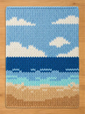 Image depicting a scenic beach with a light blue sky, white clouds, a deep blue ocean, turquoise shoreline waves, and tan sand, displayed flat on a wooden floor.