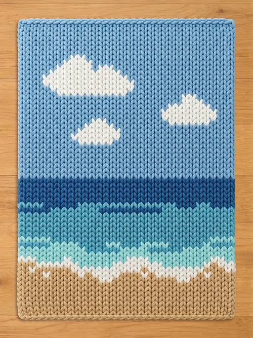 A rectangular knit blanket featuring a landscape design with a light blue sky containing white clouds, a multi-toned blue ocean with waves, and a tan sandy beach at the bottom, all constructed with a chunky, textured yarn stitch pattern.