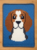 A knitted square featuring a beagle dog design. The dog's head is rendered in shades of brown, white, black, and gray, set against a vibrant blue background. The piece is crafted with a visible knit texture.