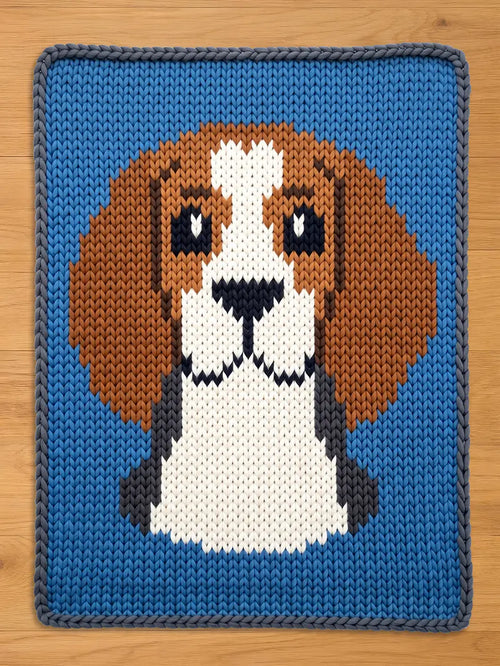A knitted square featuring a beagle dog design. The dog's head is rendered in shades of brown, white, black, and gray, set against a vibrant blue background. The piece is crafted with a visible knit texture.