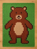 A knitted blanket featuring a pixelated brown bear with a cream-colored belly and a green background. The bear has a smiling face and is centered on the rectangular rug. The blanket is made of a thick, textured yarn.