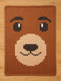 Brown knitted blanket with a bear face design. The bear's face features brown eyes with white accents, a tan muzzle, and a black nose and mouth. The blanket is rectangular and made of thick, textured yarn.