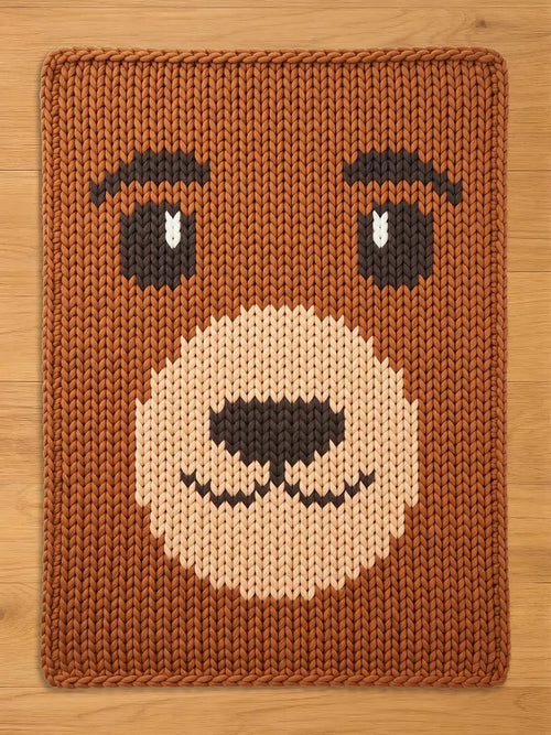 Brown knitted blanket with a bear face design. The bear's face features brown eyes with white accents, a tan muzzle, and a black nose and mouth. The blanket is rectangular and made of thick, textured yarn.