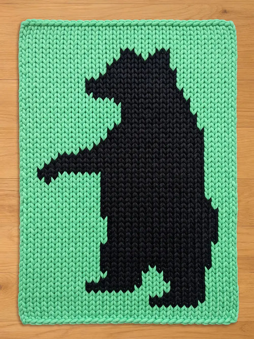 A knitted blanket featuring a black bear silhouette against a light green background. The bear is depicted in a simple, geometric style. The blanket has a rectangular shape and a textured appearance.