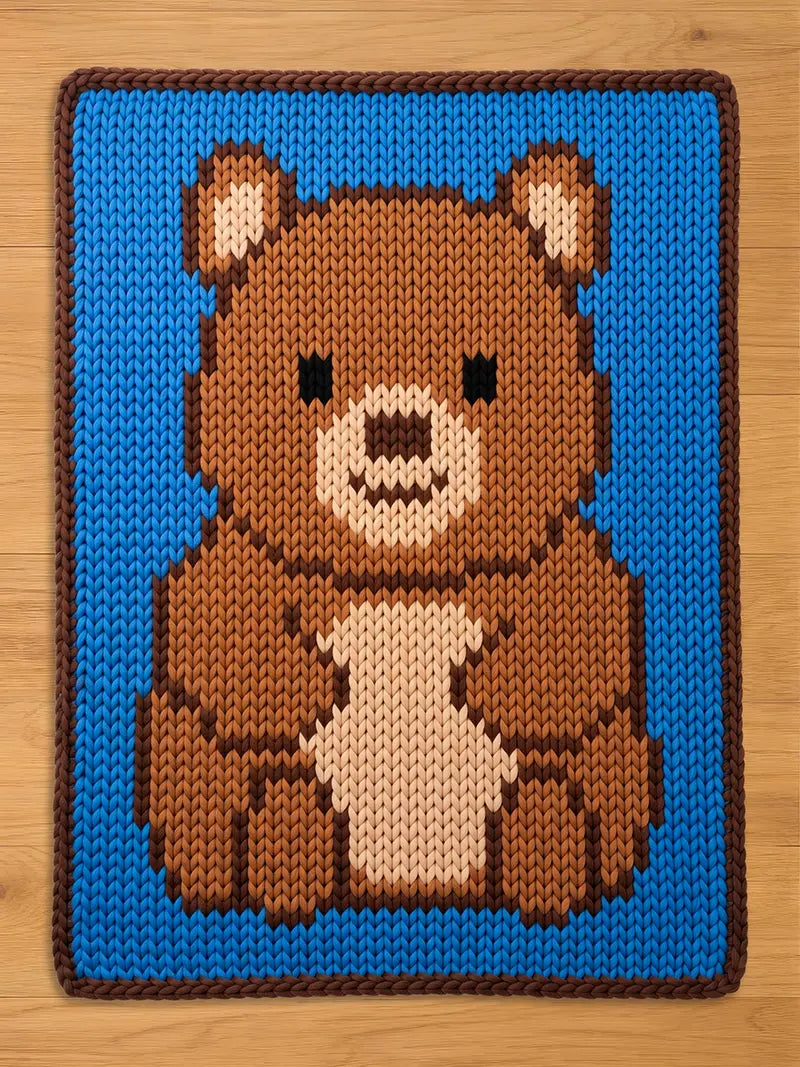 A square, knitted blanket featuring a pixelated brown bear design on a light blue background. The bear has a white chest and muzzle, with dark brown accents and black eyes. The blanket is made of a soft, textured material.