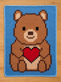 A knitted chunky blanket featuring a brown teddy bear holding a red heart. The bear has a light brown snout and ears, with black eyes. The background is a solid light blue, and the edges are also blue.