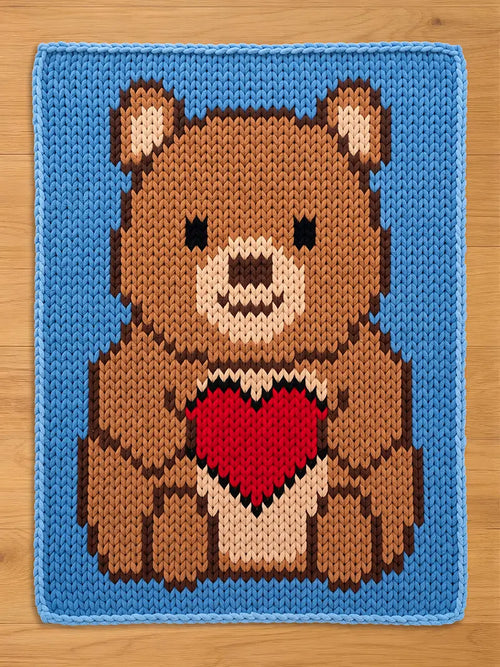 A knitted chunky blanket featuring a brown teddy bear holding a red heart. The bear has a light brown snout and ears, with black eyes. The background is a solid light blue, and the edges are also blue.