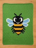 A knitted green blanket with a large, central design of a cartoon bee. The bee features a black head and body stripes, yellow stripes, and light blue wings. The blanket has a green border.